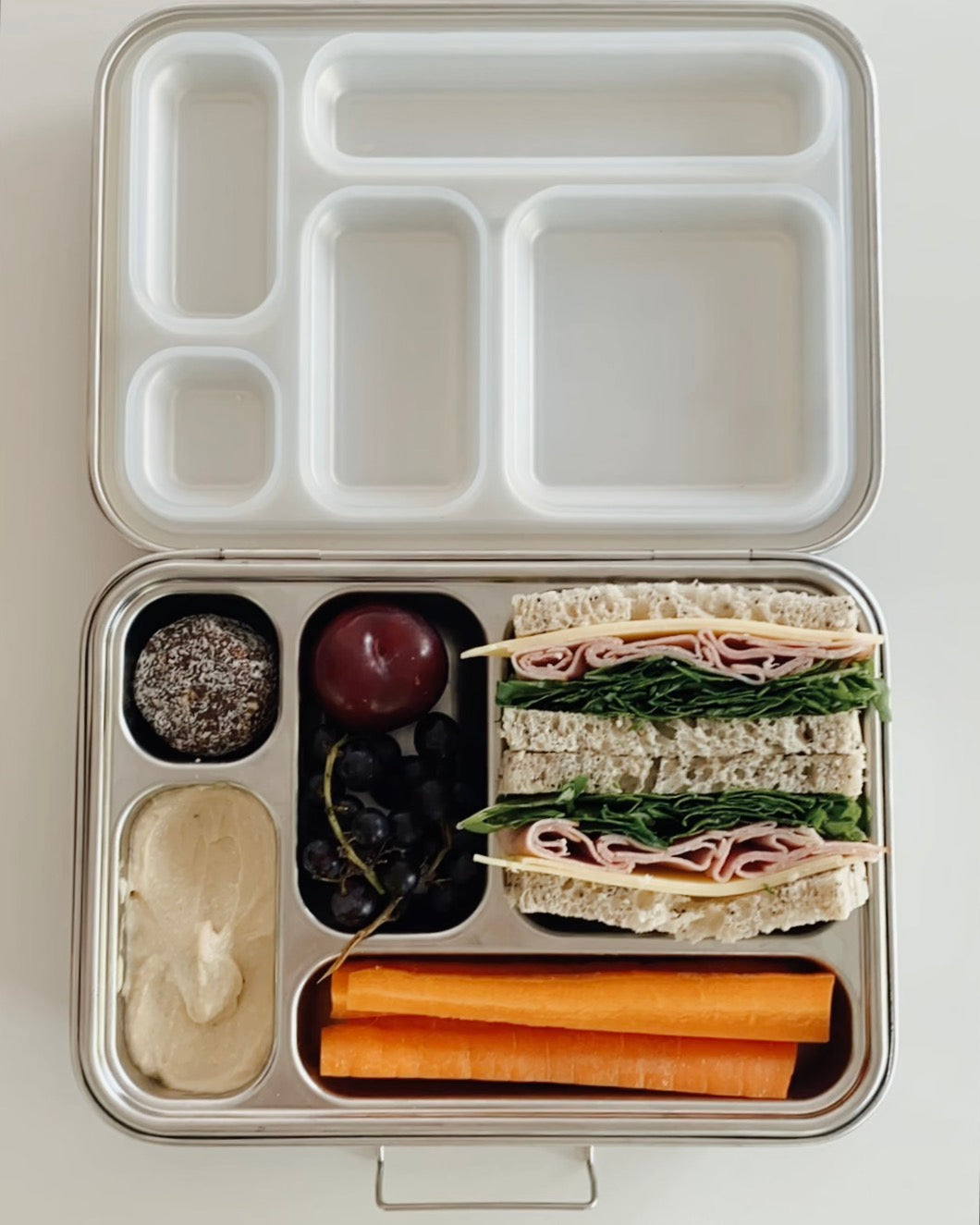 Stainless steel bento box with 5 compartments. Sustainable, eco-friendly, and zero plastic lunchbox alternative. Raw treat, carrot sticks with hummus, fruit, and ham cheese lettuce sandwich.