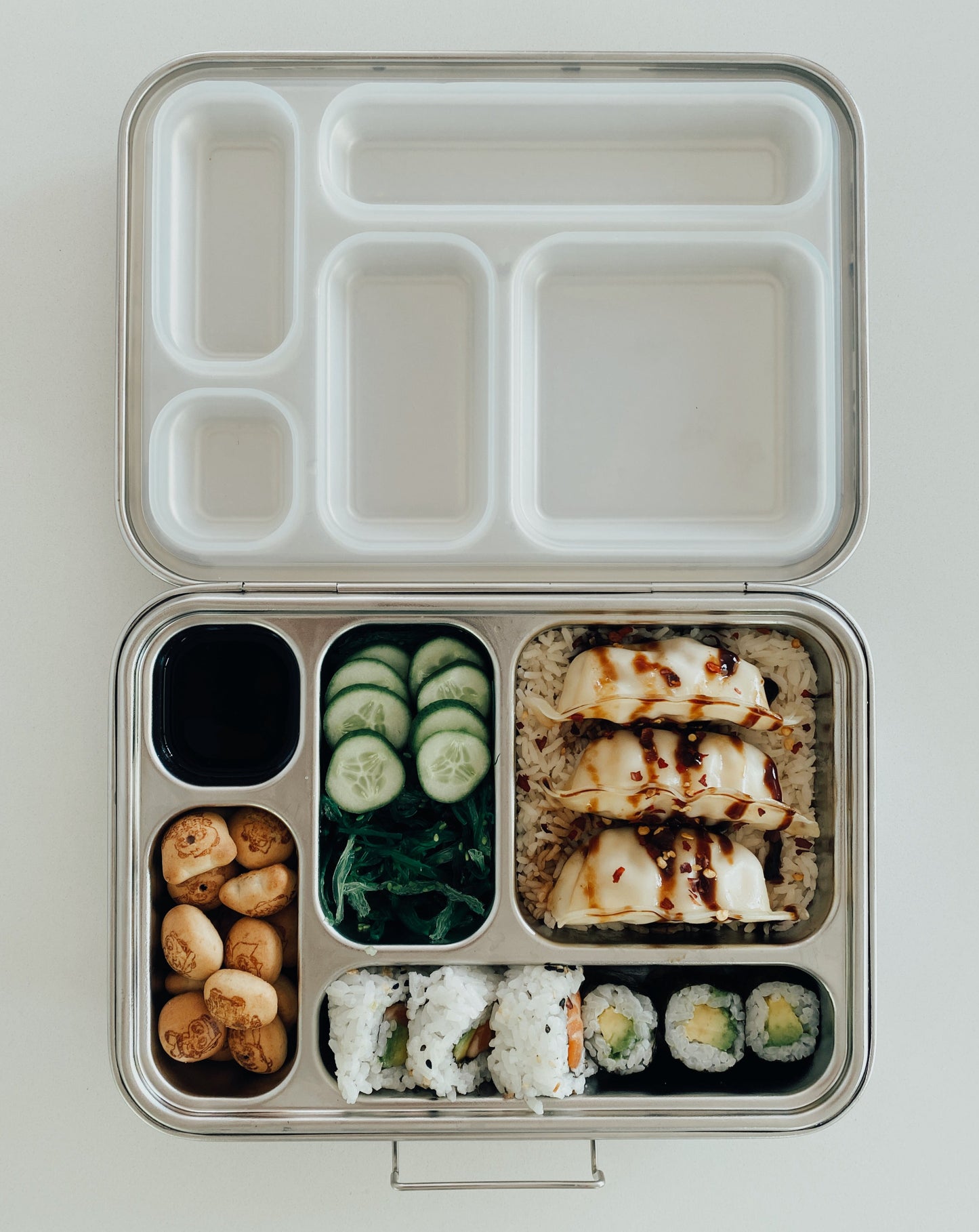 Stainless steel bento box with 5 compartments. Sustainable, eco-friendly, and zero plastic lunchbox alternative. Traditional bento arrangement with sushi, bed of rice, pork gyoza, seaweed salad, pickled cucumber, dipping soy sauce, and chocolate biscuit treats.