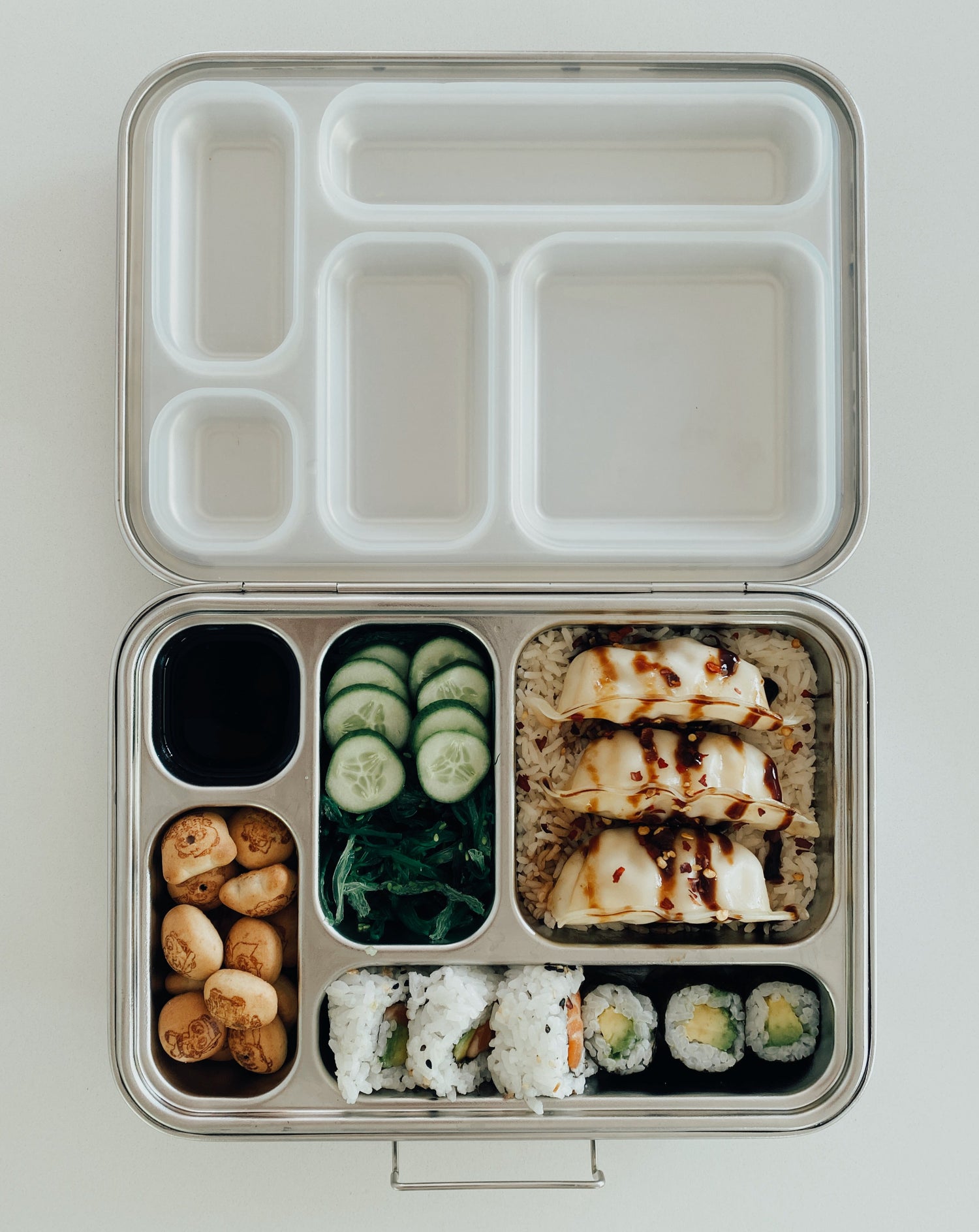 Stainless steel bento box with 5 compartments. Sustainable, eco-friendly, and zero plastic lunchbox alternative. Traditional bento arrangement with sushi, bed of rice, pork gyoza, seaweed salad, pickled cucumber, dipping soy sauce, and chocolate biscuit treats.