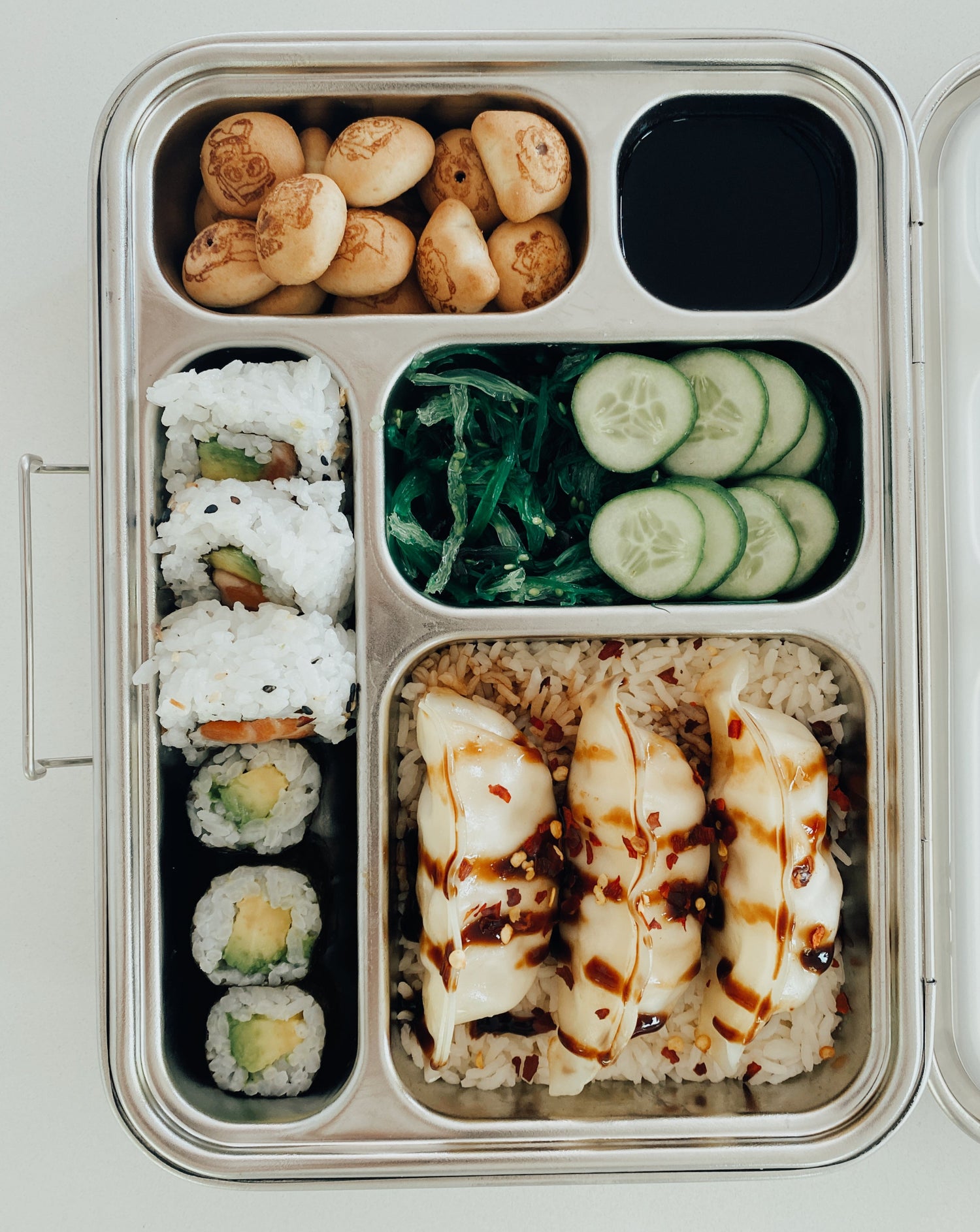 Stainless steel bento box with 5 compartments. Sustainable, eco-friendly, and zero plastic lunchbox alternative. Traditional bento arrangement with sushi, bed of rice, pork gyoza, seaweed salad, pickled cucumber, dipping soy sauce, and chocolate biscuit treats. 