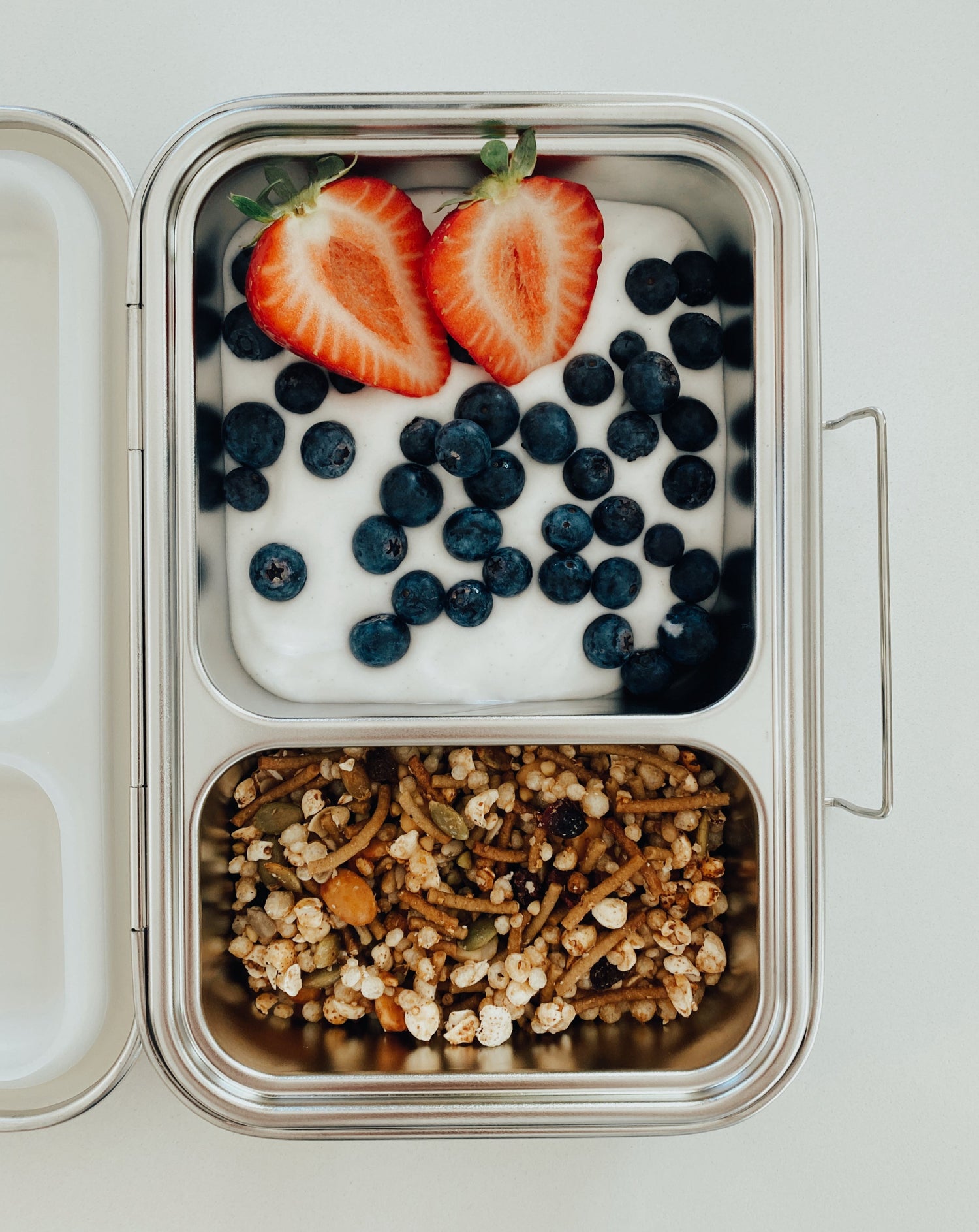 Stainless steel bento box with 2 compartments respectively. Sustainable, eco-friendly, and zero plastic lunchbox alternative. Breakfast is served with coconut yogurt, blueberries, strawberries, and gluten-free granola.