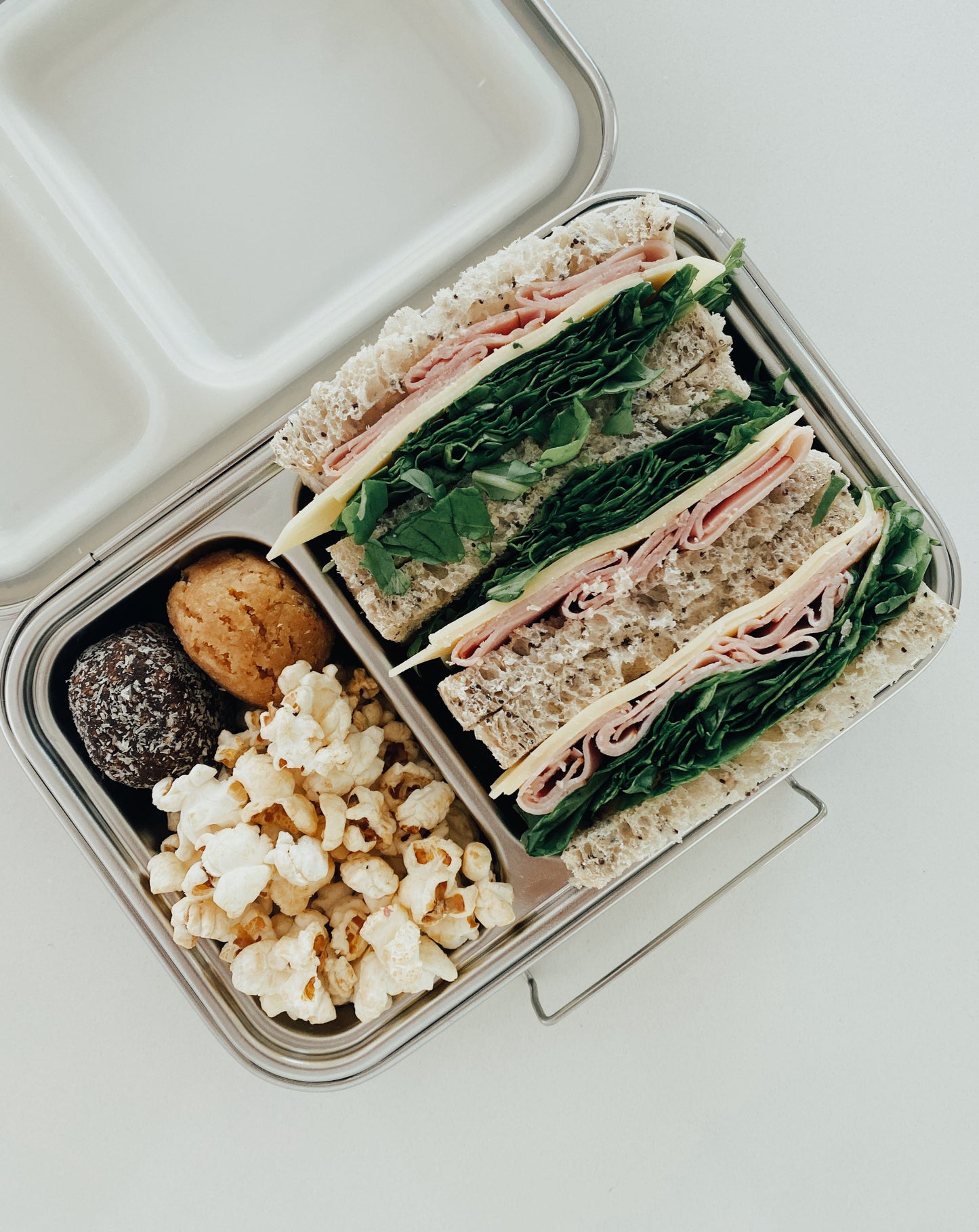 Stainless steel bento box with 5 compartments. Sustainable, eco-friendly, and zero plastic lunchbox alternative. Raw treat, carrot sticks with hummus, fruit, and ham cheese lettuce sandwich.